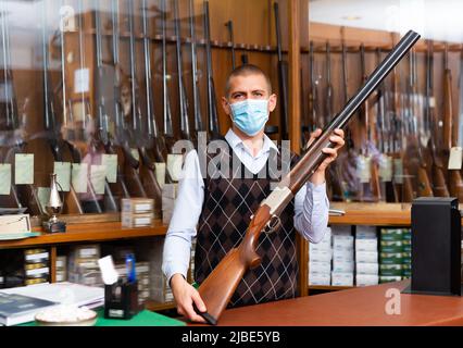 Venditore in maschera medica mostrando doppio fucile da caccia barreled Foto Stock