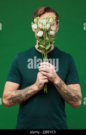 Ritratto verticale dell'uomo tatuato che tiene rose bianche e copre il volto su sfondo verde vibrante Foto Stock