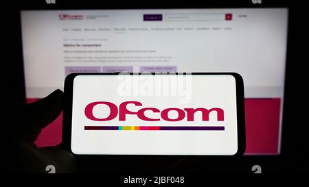 Persona che tiene sullo schermo il cellulare con il logo dell'ufficio delle comunicazioni dell'autorità britannica (UFCOM) davanti alla pagina web. Mettere a fuoco sul display del telefono. Foto Stock