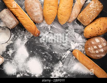 un vasto assortimento di diversi tipi di pane disposti su sfondo nero con farina, dolci caldi freschi, farina di grano, produzione di pane Foto Stock