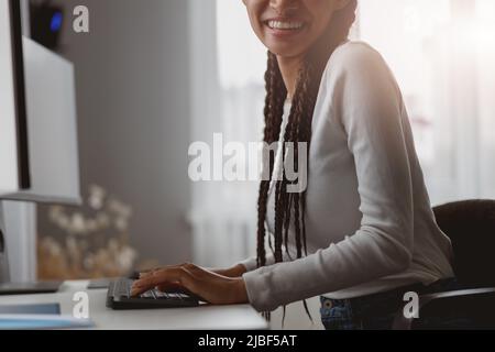 Primo piano di una giovane donna sorridente seduta alla scrivania in appartamento che lavora al computer Foto Stock