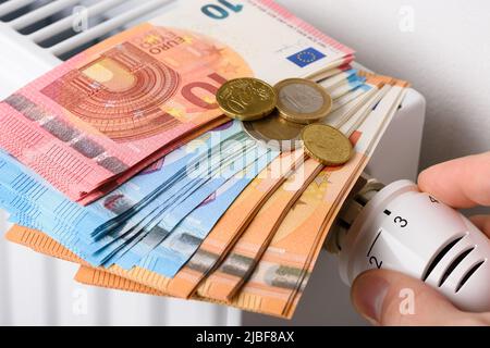 Regolazione manuale della manopola della valvola del termostato di temperatura del radiatore di riscaldamento con pila di banconote in euro. Costi di riscaldamento costosi e in aumento Foto Stock
