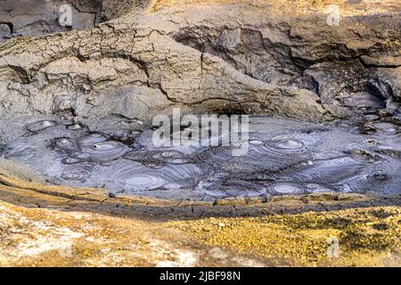 Motivi circolari di bolle coprono la superficie di una pozza di fango grigia nell'area geotermica di Hverir ai piedi del monte Námafjall, a est del lago Mývatn nel nord-est dell'Islanda, incorniciata da roccia grigia pallida e crepata. Hverir si trova al passo di Námaskarð, appartiene al sistema vulcanico di Krafla ed è nota per le sue pozze di fango gorgoglianti e le fumarole fumanti che rilasciano gas solforoso. Foto Stock
