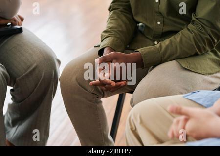 Mani afflitto di giovane afroamericano travagliato in casualwear seduto tra le altre persone durante la sessione psicologica Foto Stock