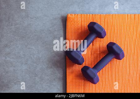 Vista dall'alto dei manubri viola e del tappeto arancione posto sul pavimento grigio durante l'allenamento fitness Foto Stock