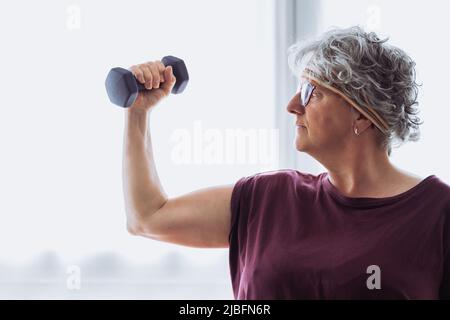 Donna anziana potente con i capelli corti grigio che sollevano il dumbbell e che mostra il bicep contro la finestra durante l'allenamento di idoneità al mattino a casa Foto Stock