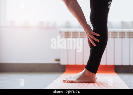 Anonimo donna a piedi nudi nei leggings che toccano la gamba e si piegano in avanti mentre si levano in piedi sul tappetino durante l'allenamento di fitness a casa Foto Stock