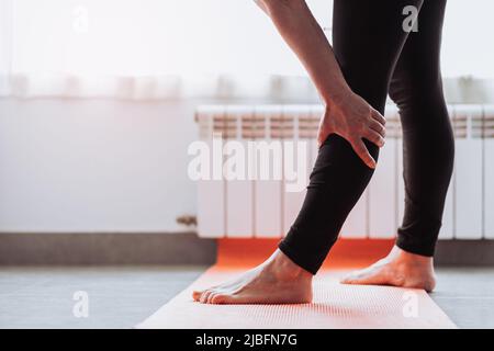 Anonimo donna a piedi nudi nei leggings che toccano la gamba e si piegano in avanti mentre si levano in piedi sul tappetino durante l'allenamento di fitness a casa Foto Stock