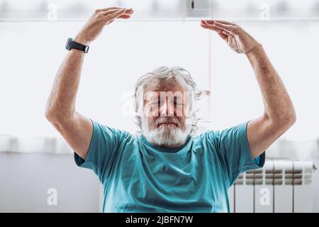 Uomo anziano in t shirt blu che alza le braccia e medita con occhi chiusi contro la finestra durante la sessione di yoga al mattino a casa Foto Stock