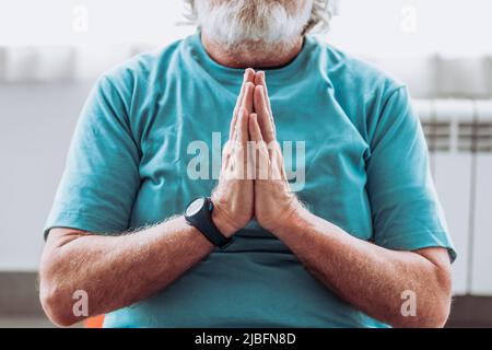 Calmo corto irriconoscibile uomo anziano in activewear che stringe le mani e meditating mentre si siede durante la sessione di yoga nella stanza luminosa Foto Stock