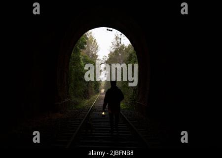 Silhouette di turista anonimo con lampada a piedi su rotaie e ammirare la foresta in tunnel scuro in campagna Foto Stock