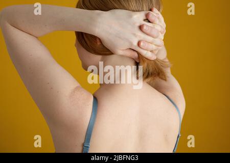 Vista posteriore di una donna irriconoscibile con capelli zenzero e corpo curvo in piedi contro il muro giallo in casa Foto Stock
