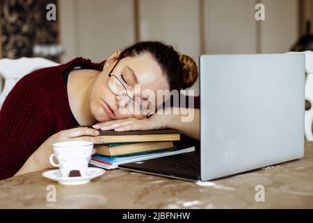 Ritratto di esausta donna di mezza età seduta a tavola vicino a tazza, laptop, dormire su braccia incrociate su mucchio di libri. Foto Stock