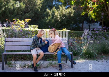 Bambini felici seduti su panca all'aperto nel parco, parlare e preparare i compiti. Foto Stock