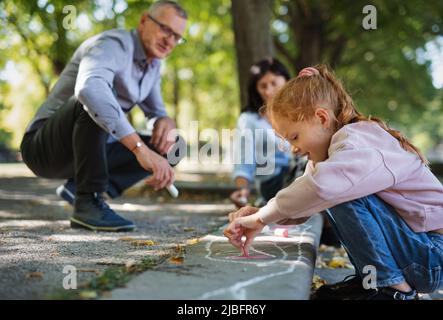 Coppia senior con nipoti che disegnano con gessi su marciapiedi all'aperto nel parco. Foto Stock