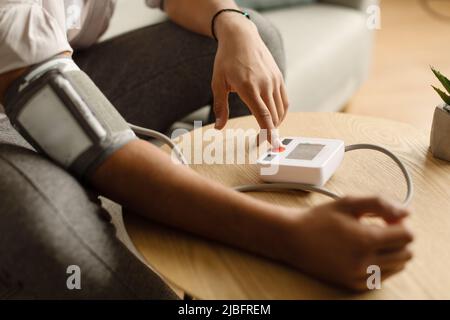 Vista ritagliata dell'uomo nero millenario seduto sul divano a casa, controllando la pressione sanguigna, utilizzando un moderno tonometro Foto Stock