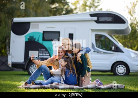 Una giovane famiglia felice con due bambini che prendono selfie con caravan in background all'aperto. Foto Stock