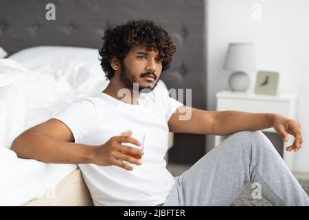 Ragazzo orientale sconvolto che beve alcol, seduto al piano a letto Foto Stock