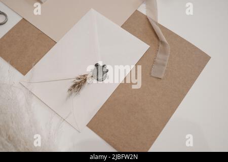 Vista dall'alto della composizione di fogli di carta bianchi, marroni, rosa, busta bianca sigillata con cera grigia e ramoscello essiccato. Foto Stock
