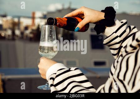 Donna che indossa una felpa a righe, nera scricchiolante a portata di mano, versando champagne dalla bottiglia al bicchiere sullo sfondo della città. Foto Stock