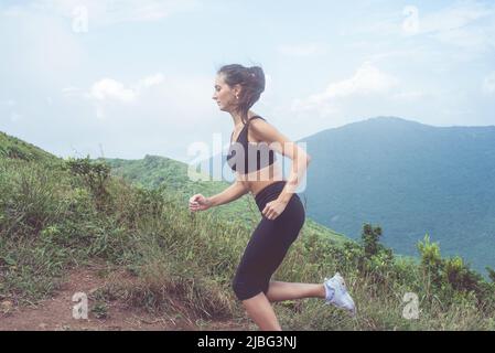 Vista laterale dell'atleta femminile in allenamento, corsa all'aperto in montagne verdi. Giovane donna che indossa abbigliamento sportivo nero che si allena all'aperto in estate. Foto Stock