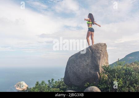 Vista posteriore di sportivo ispirato in piedi su una roccia in montagna con le braccia distese ammirando la vista del cielo nuvoloso e del mare in estate. Foto Stock