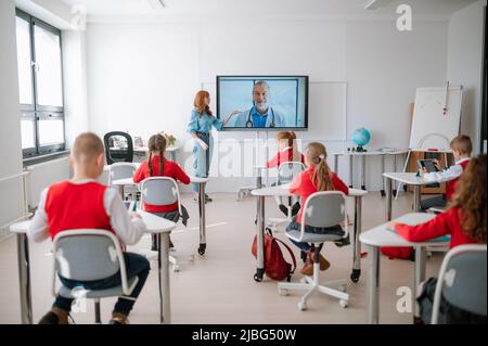 Vista posteriore, classe di studenti seduti, ascoltare l'insegnante e wathing online lezione con il medico in aula Foto Stock