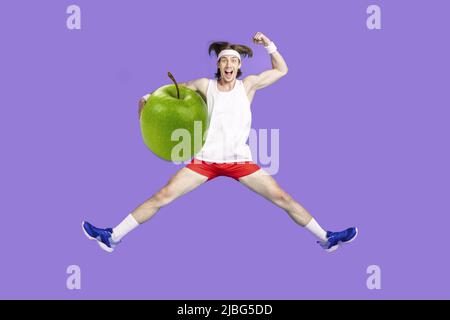 Ragazzo pazzo overjoyed che salta la mela gigante alta tenuta hanno spuntino prima di addestramento che si riscalda allungando su sfondo viola Foto Stock