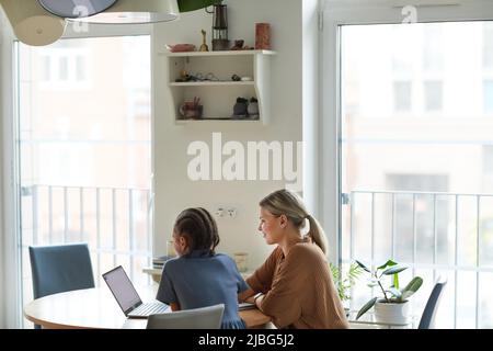 Minimo scatto di famiglia multietnica, madre e figlia guardando lo schermo del laptop mentre studiano insieme a casa in un ambiente minimo, spazio di copia Foto Stock