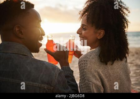 Sorridente giovane coppia multietnica bere succo su una spiaggia al tramonto Foto Stock
