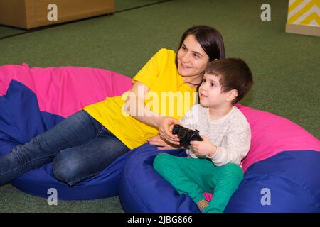 Madre e figlio giocano nella console di gioco, il ragazzo tiene il joystick, guardando lateralmente nella sala giochi per i bambini. Foto Stock
