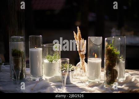 Banchetti per matrimoni all'aperto nella foresta. Tavolo da pranzo decorato in stile boho con candele, stoffa bianca, fiori, servito con piatti, stoviglie Foto Stock