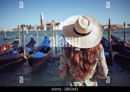 Vista da dietro elegante viaggiatore solista donna in abito floreale con cappello sul terrapieno a Venezia, Italia. Foto Stock