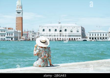 Vista da dietro l'elegante viaggiatore solista donna in abito floreale con cappello seduto sul lungomare sull'isola di San Giorgio maggiore. Foto Stock