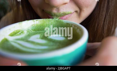 Donna drink alla moda matcha latte con arte in caffè e schiuma verde rimane sulle labbra. Giovane donna lecca la schiuma matcha verde dal primo piano delle labbra Foto Stock