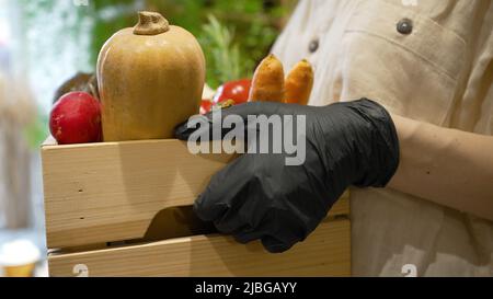Donna in guanti di gomma nera porta scatola di legno piena di verdure fresche. Il lavoratore della cucina del caffè trasporta la scatola con i closeup organici delle verdure Foto Stock