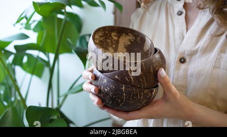 La giovane donna tiene piccole ciotole vuote marroni nelle mani con manicure bianco contro la pianta verde. La donna si affaccia su nuovi piatti per il primo piano del caffè Foto Stock