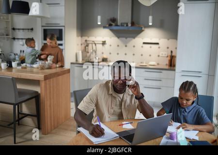 Vista frontale del padre nero che lavora da casa con la figlia seduta al tavolo accanto a lui, spazio copia Foto Stock