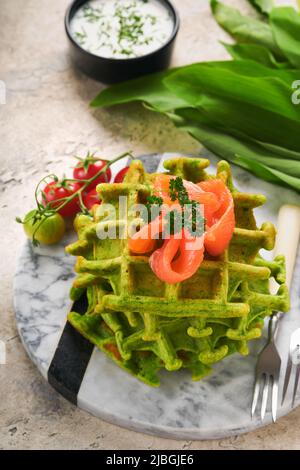 Waffle belgi verdi. Spinaci o aglio selvatico o pesto cialde con salmone rosso e salsa di panna su sfondo grigio tavolo di cemento. Deliziosa colazione, Foto Stock