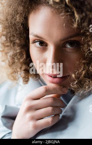 primo piano ritratto di affascinante donna ricci con occhi marroni che toccano il mento e guardando la macchina fotografica Foto Stock