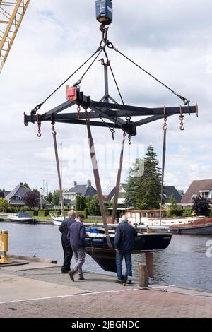 La barca a vela, sospesa con pneumatici su una costruzione in acciaio, viene sollevata dal molo in acqua con una gru. La gente accompagna la barca Foto Stock