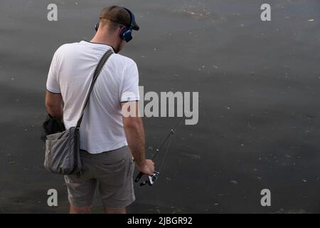 Il giovane uomo in camicia bianca, berretto, cuffie e borsa a spalla è pesca con una canna da pesca Foto Stock