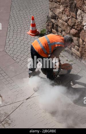 Uomo in giacca di sicurezza arancione fa fessure strette con un macinacaffè in una strada di pietra. La grinta e la polvere gli spolverano intorno. Concentrarsi sui capelli del lavoratore Foto Stock