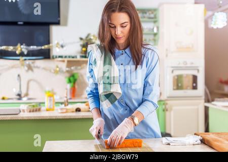 Cuoca femminile che lavora in guanti facendo rotoli di sushi giapponesi che li affettano su un tappeto di bambù in piedi in cucina. Foto Stock