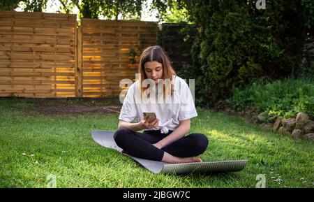 Una giovane donna si siede su un tappetino per il fitness e guarda lo schermo dello smartphone. Foto Stock