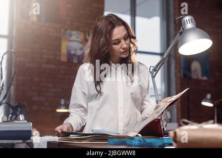 Giovane donna dressmaker scelta del materiale dal catalogo in studio. Personalizza l'aspetto attraverso i tessuti mentre sei in piedi in un'officina di cucito. Foto Stock