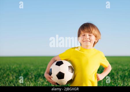 Il bambino felice finge di essere un giocatore di calcio. Ritratto di bambino all'aperto. Ragazzo contro campo verde. Successo e concetto del vincitore Foto Stock