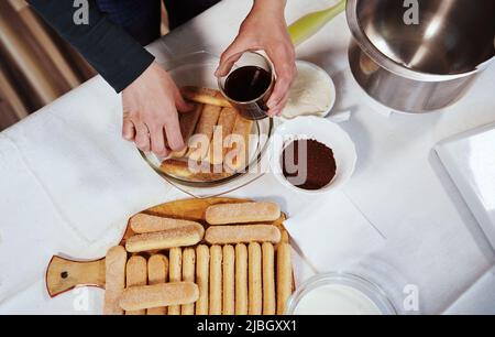 Vista dall'alto verso le mani femminili che impilano biscotti salati immersi in un caffè espresso dolce forte su un piatto mentre si prepara la torta Tiramisu fatta in casa Foto Stock