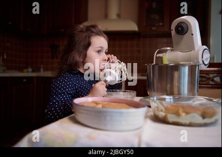 Adorabile caucasica bambina lecca sussurrare con la panna montata dolce rimanente mentre la mamma sta preparando il dessert tiramisù in cucina a casa Foto Stock