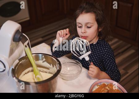 Vista dall'alto di una adorabile bambina europea che lecca la deliziosa panna montata dal mixer del robot da cucina mentre sua madre è assente Foto Stock